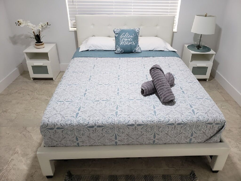 Un lit bien fait, avec une couvre-lit à motifs, un oreiller orné d’une citation inspirante et une serviette enroulée posée sur le lit.