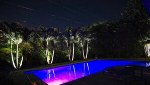 A nighttime swimming pool with illuminated palm trees and lounge chairs. - Blue Tempo - Steps to Wilton Dr, Heated Salt Pool, Huge Yard (Wilton Manors)