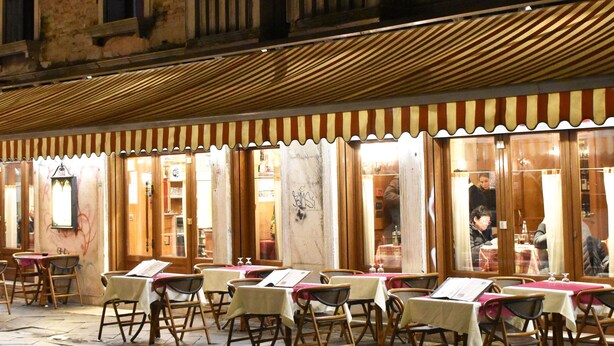 An outdoor cafe with tables and chairs, a wooden pergola, and a building with arched windows in the background. - Veniceluxury (Venice)