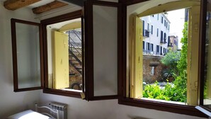 A bedroom with a bed, a bedside table, a radiator, and a window with a view of buildings and greenery outside. - Veniceluxury (Venice)
