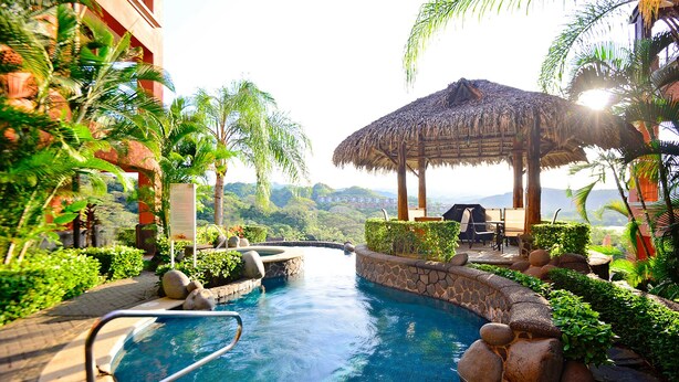 A pool area with a thatched gazebo, surrounded by lush greenery and a stone wall. - Luxury Ground Floor  Condo, close to pool access to beach club at Los Suenos. (PUNTARENAS)