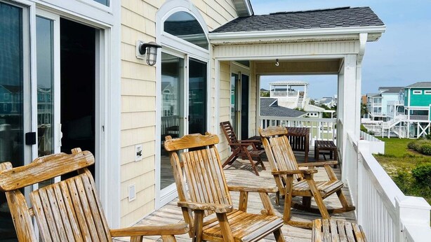 Um deck de madeira com cadeiras e uma mesa, área coberta com guarda-corpo, e vista para casas ao fundo.