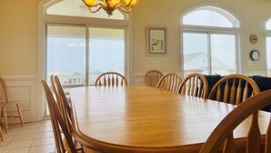 Uma sala de jantar com uma grande mesa de madeira, seis cadeiras, um candelabro, janelas amplas e vista para o exterior.