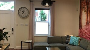 A living room with a sectional sofa, a glass coffee table, a wicker chair, a patterned rug, a wall clock, a window with curtains, and recessed ceiling lighting.