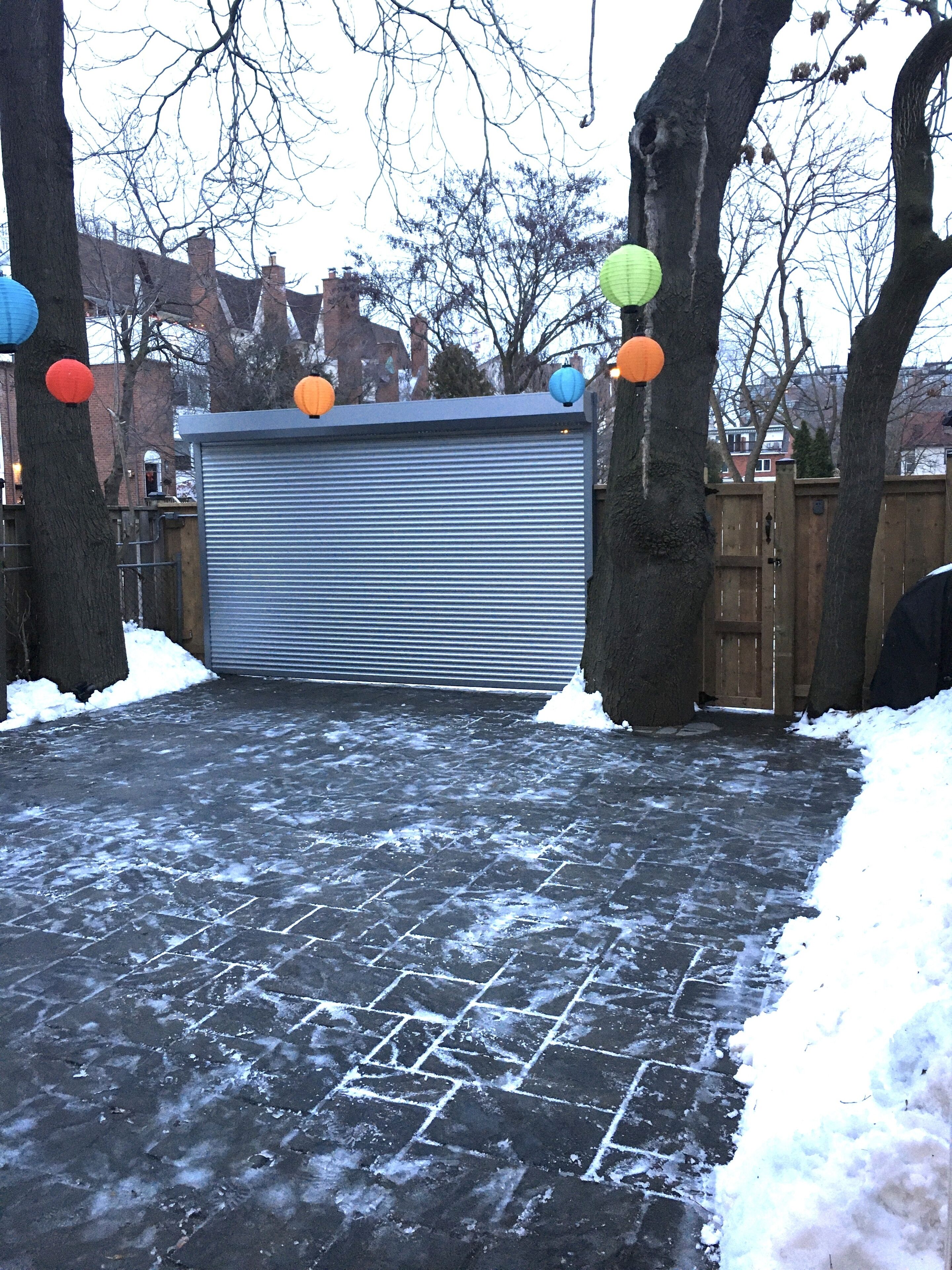 A garage with a closed metal door, surrounded by trees and a wooden fence. Snow is visible on the ground and on the fence.