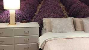 A bedroom with a lavender field mural on the wall, a bedside lamp, a white dresser, a patterned rug, and a bed with floral bedding.