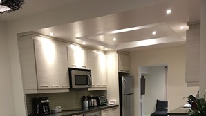 A modern kitchen with white cabinets, a microwave, a coffee maker, and a refrigerator. There is a dining area with a glass table and chairs, and a living room with a sofa and a coffee table.