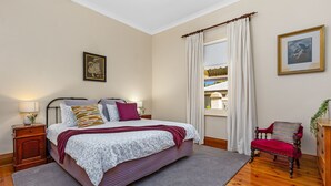 A bedroom with a bed, a chair, a nightstand, a window with curtains, and a framed picture on the wall. - Miriam's Cottage Self Contained 2 bedroom cottage (Tanunda)