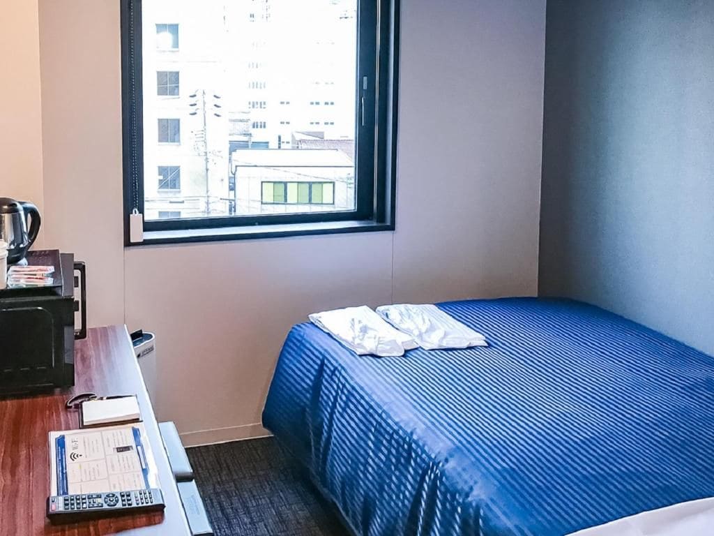 Une chambre d’hôtel avec un lit, un bureau équipé d’un ordinateur et une fenêtre donnant sur des immeubles.