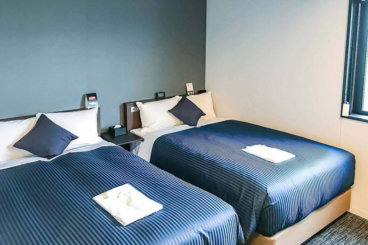 Hotelkamer met twee bedden, elk voorzien van een blauwe sprei en kussens, een nachtkastje met een lamp en een raam met gordijnen.