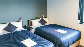 A hotel room with two beds, each with a blue bedspread and pillows, a nightstand with a lamp, and a window with curtains.