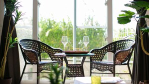 A room with wicker chairs and a table, two wine glasses on the table, and a view of greenery outside. - Irie House (Onna)