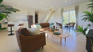 A spacious living area with a sofa, coffee table, and chairs. A kitchen is visible in the background with a staircase leading to an upper level. - Irie House (Onna)
