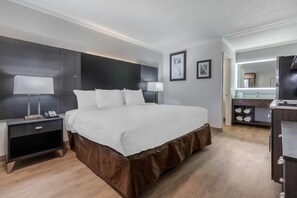 A hotel room with a large bed, bedside tables, a desk, and a mirror. - Suburban Studios at the University (Conway)