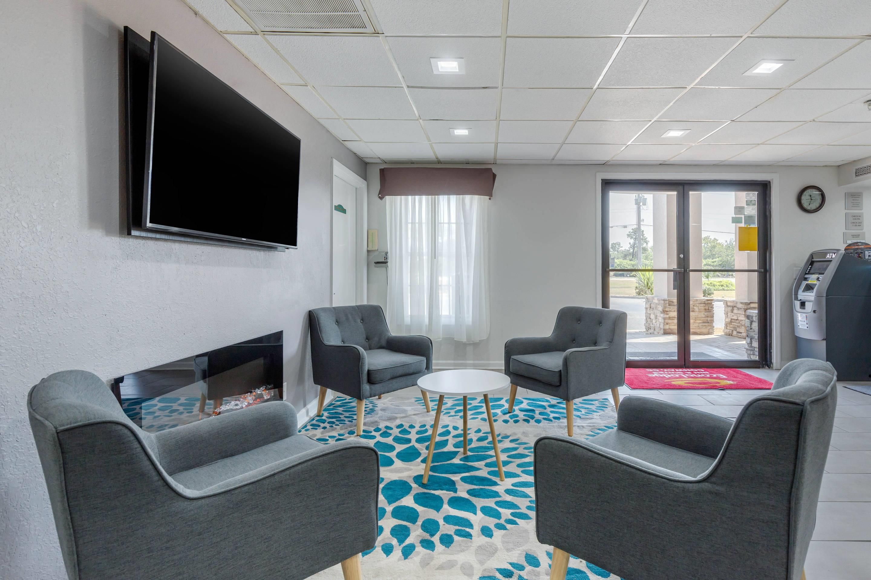 Habitación de hotel con televisor de pantalla plana montado en la pared, dos sillones grises, una mesita redonda, una chimenea y vistas al exterior a través de una puerta de cristal.