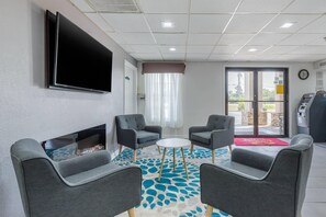 A hotel room with a flat-screen TV mounted on the wall, two grey armchairs, a small round table, a fireplace, and a view of the outside through a glass door. - Suburban Studios at the University (Conway)