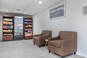 Un área de espera con máquinas expendedoras llenas de aperitivos y bebidas, dos sillones marrones y un cuadro en la pared.