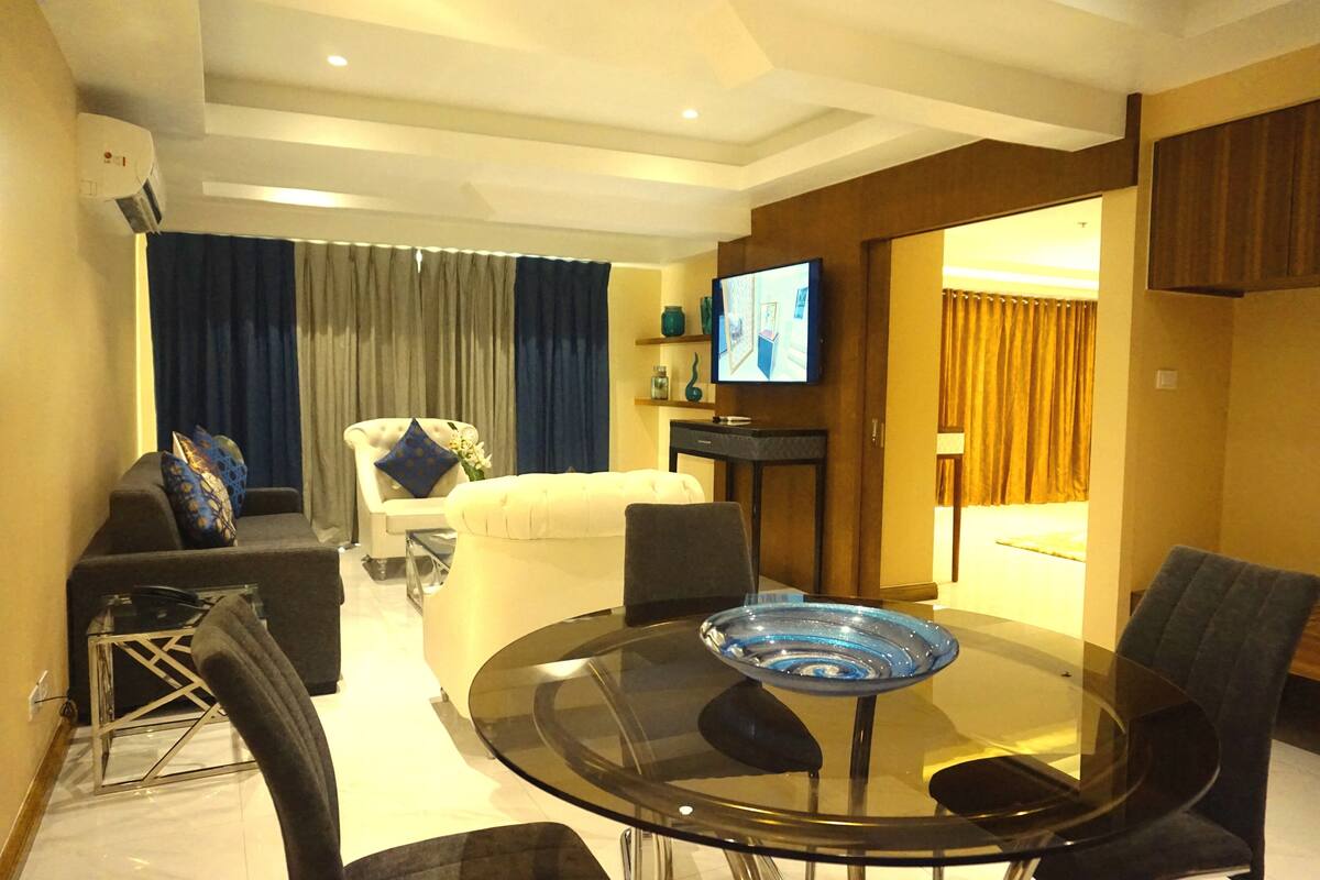 A modern living room with a glass dining table, a sofa, a white armchair, and a television mounted on the wall.