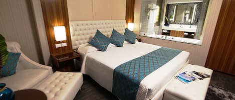 A hotel room with a large bed, a sitting area with a chair and ottoman, a nightstand, a mirror, and a window with a view.