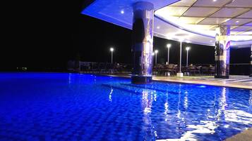Una piscina iluminada con luces azules en la noche, rodeada de sillones de descanso y un área de estar techada.