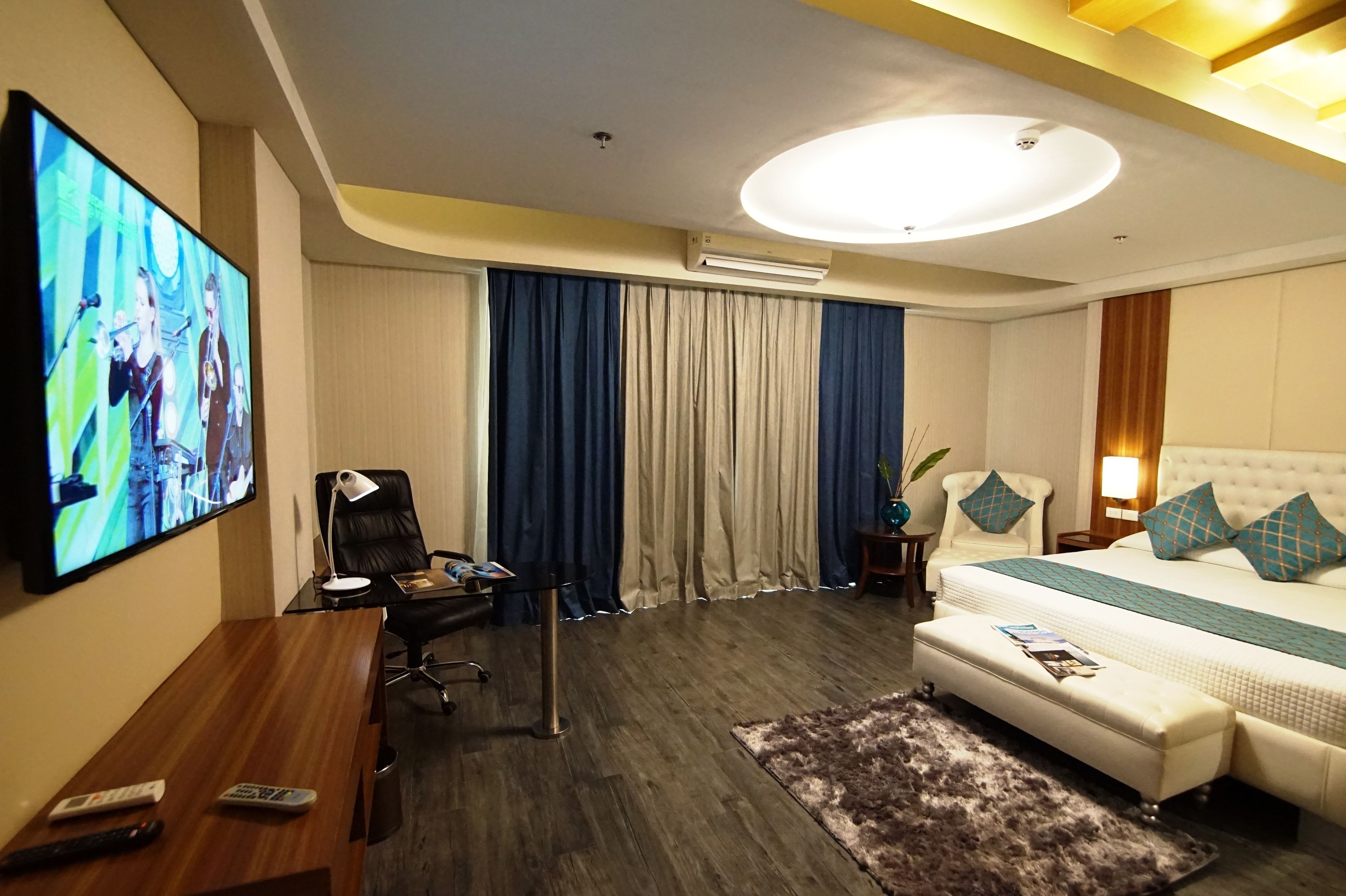 A hotel room with a large flat-screen TV, a desk with a chair, a bed with a white headboard, and a large window with blue curtains.