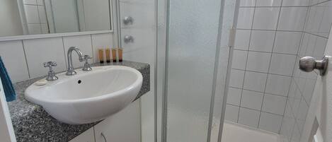 Ein Badezimmer mit weißem Waschbecken, einem Spiegel, einer Duschkabine und dunkel gefliestem Boden.