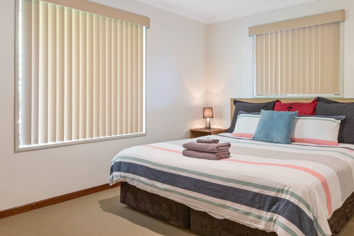 A bedroom with a bed, pillows, a nightstand with a lamp, and large windows with blinds.
