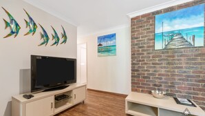A modern living room with a flat-screen TV on a white cabinet, a brick accent wall with a painting of a pier, and decorative fish wall art. - Mandurah Caravan & Tourist Park (Furnissdale)