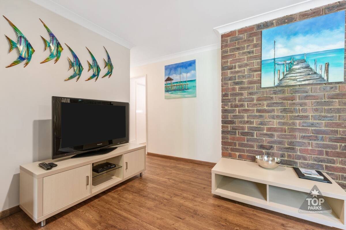 A modern living room with a flat-screen TV on a white cabinet, a brick accent wall with a painting of a pier, and decorative fish wall art.