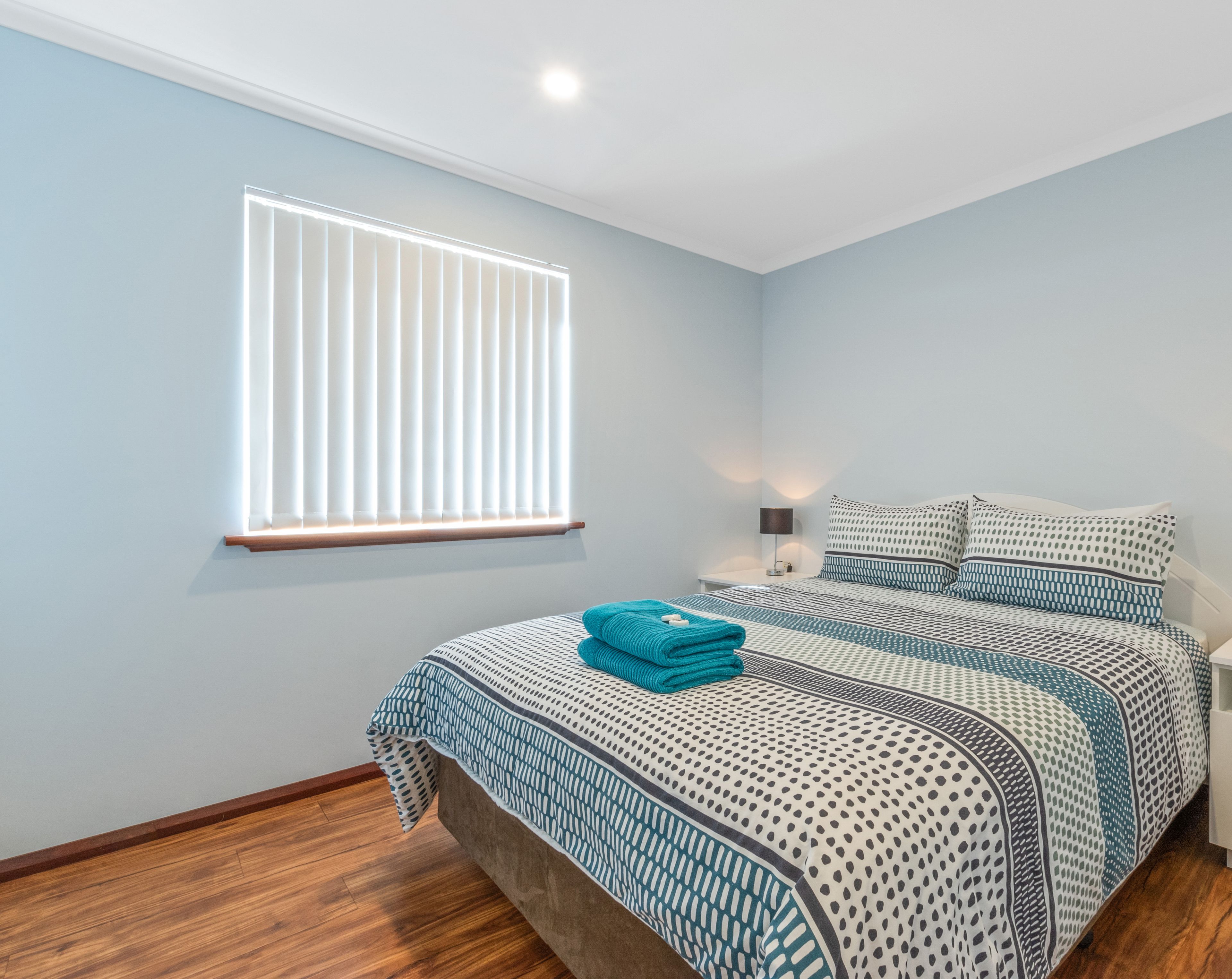 A bedroom with a bed, a window with blinds, a nightstand with a lamp, and a wooden floor.