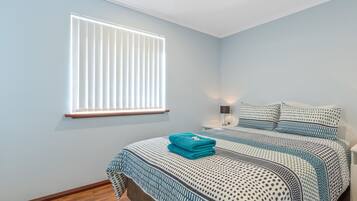 A bedroom with a bed, a window with blinds, a nightstand with a lamp, and a wooden floor.