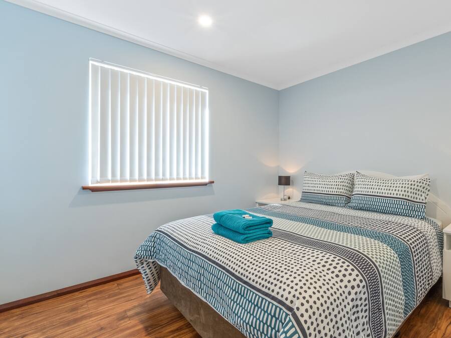 A bedroom with a bed, a window with blinds, a nightstand with a lamp, and a wooden floor.