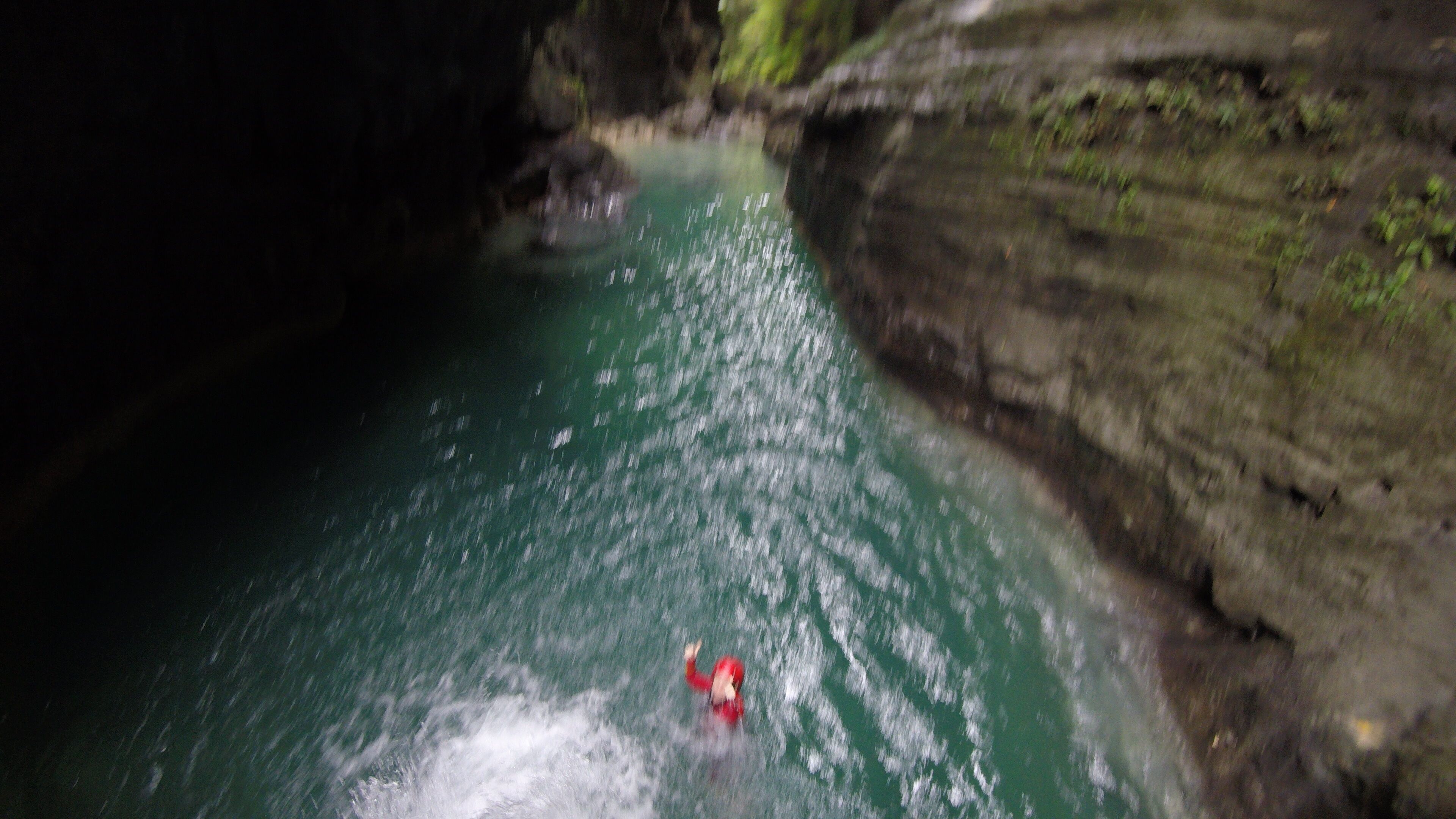A person in a red jacket swimming in a river surrounded by rocky cliffs.