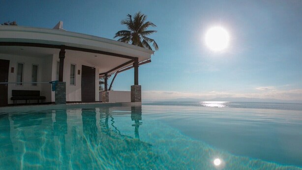 Una piscina infinita moderna con vista despejada al océano, un espacio exterior cubierto y una palmera.