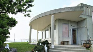 A modern building with a curved entrance, steps leading up to a door, and a sign with Japanese characters. - Minshuku Tantawan (Motobu)