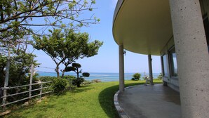 A covered outdoor area with a view of the sea, a concrete structure, and a grassy area with trees. - Minshuku Tantawan (Motobu)