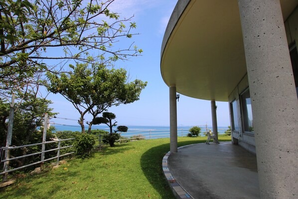 A covered outdoor area with a view of the sea, a concrete structure, and a grassy area with trees. - Minshuku Tantawan (Motobu)