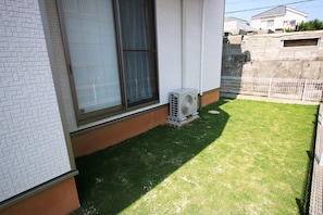 A residential backyard with a white brick wall, a window with a curtain, and a small air conditioning unit. - Double Smile (Yomitan)