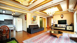 Uma sala de estar ampla com um sofá, uma mesa de centro e uma televisão. Dá para ver uma cozinha ao fundo, com frigorífico e armários.