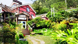Um edifício tradicional com fachada vermelha, entrada frontal com degraus e área de jardim com diversas plantas.