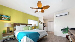 Un dormitorio con cama, mesitas de noche, ventilador de techo, unidad de aire acondicionado y ventana.