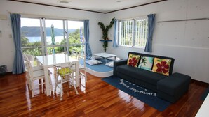 Una sala de estar con piso de madera, un sofá negro, una mesa blanca y un juego de comedor azul. Hay ventanas grandes con cortinas, una lámpara de techo y vistas al follaje exterior.