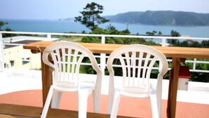Una mesa de madera con dos sillas blancas de plástico en un balcón que da a un paisaje costero con vistas al mar y colinas lejanas.