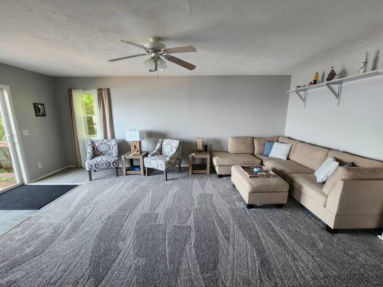 A spacious living room with a grey carpet, a ceiling fan, a beige sofa, two patterned armchairs, a coffee table, and a sideboard with decorative items.
