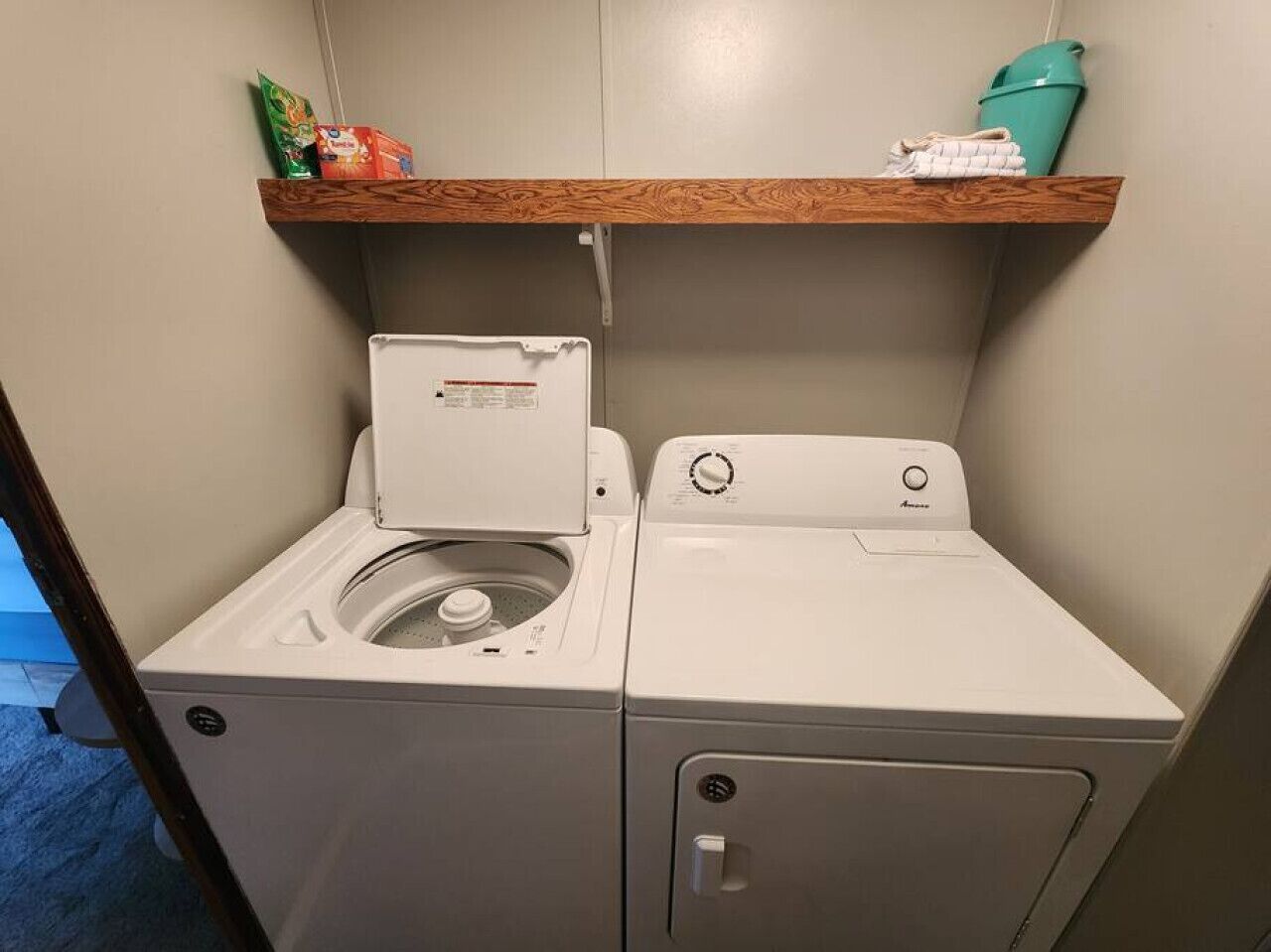 Ein kleiner Waschraum mit Waschmaschine und Trockner, einem Holzregal darüber und einem Teppichboden in Blau.