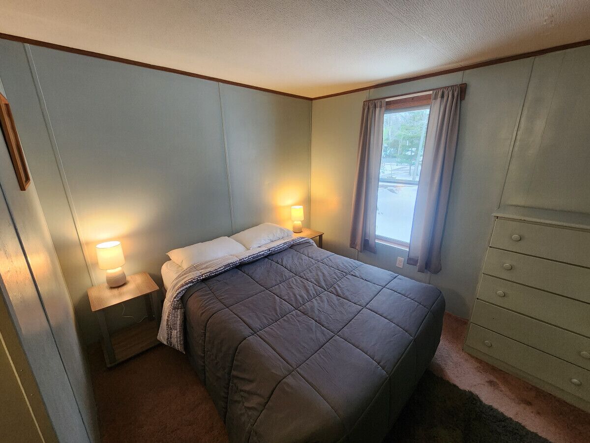 Une petite chambre à coucher, bien rangée, avec un lit, des lampes de chevet, une commode et une fenêtre avec des rideaux.