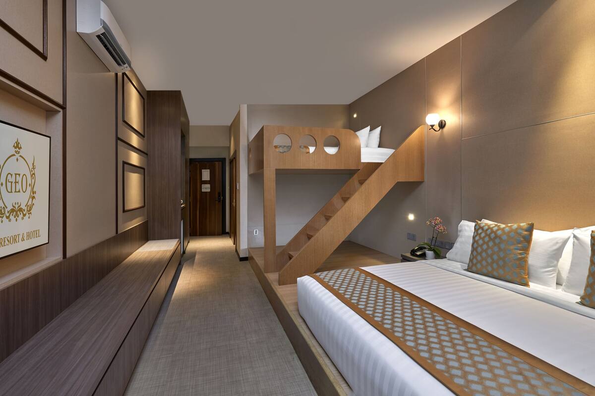 A hotel room with a bunk bed, a large bed with patterned bedding, a wooden staircase, and a wall-mounted air conditioner.