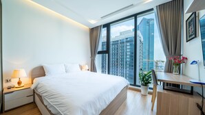A modern hotel room with a large bed, a desk, a chair, a potted plant, and a view of tall buildings. - Jaan Apartment 29 Lieu Giai (Hanoi)