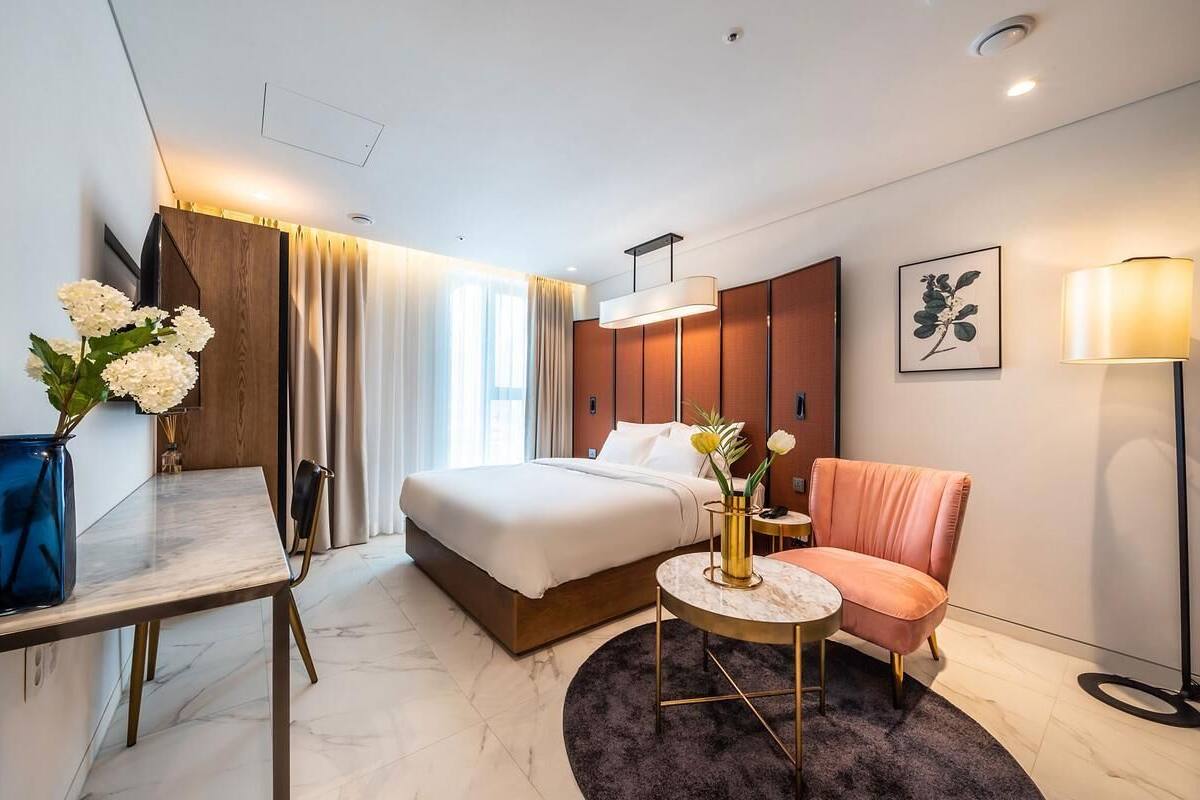 A hotel room with a bed, a desk, a chair, a round table, a floor lamp, and a vase with flowers.