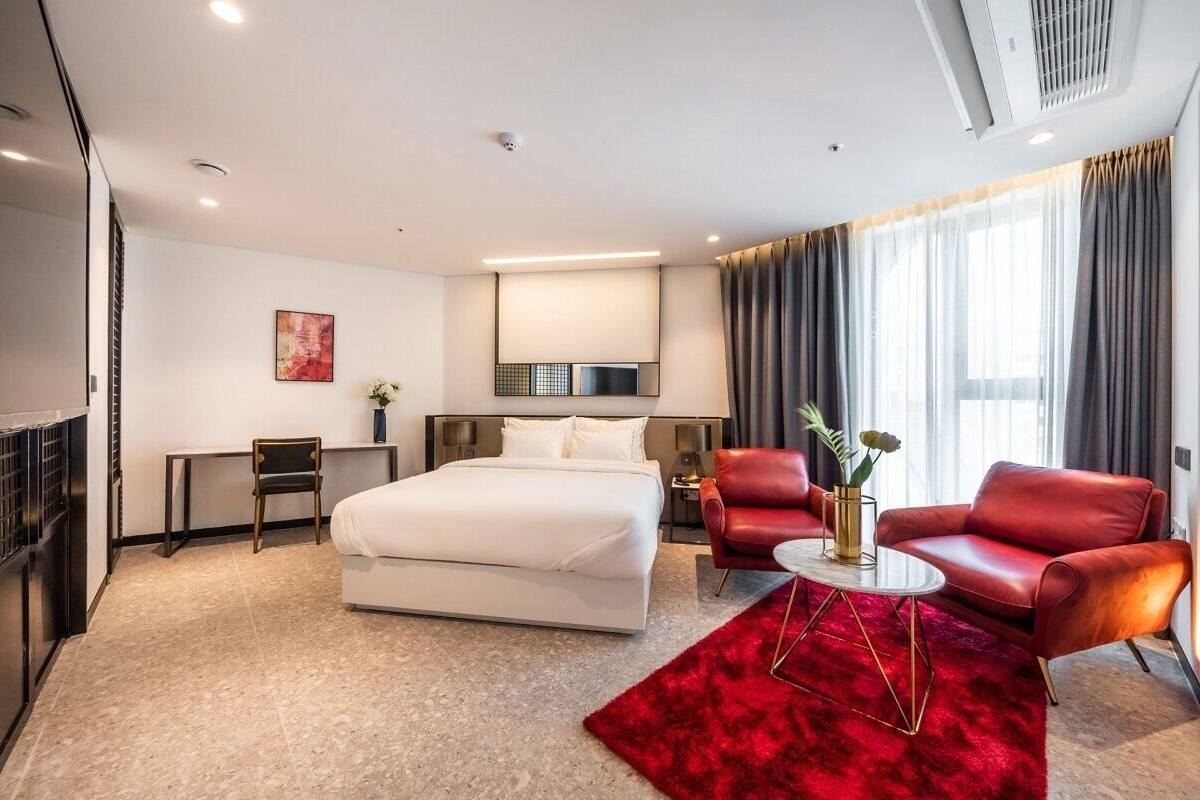 A modern hotel room with a bed, a desk, two red leather chairs, a red rug, and a large window with curtains.
